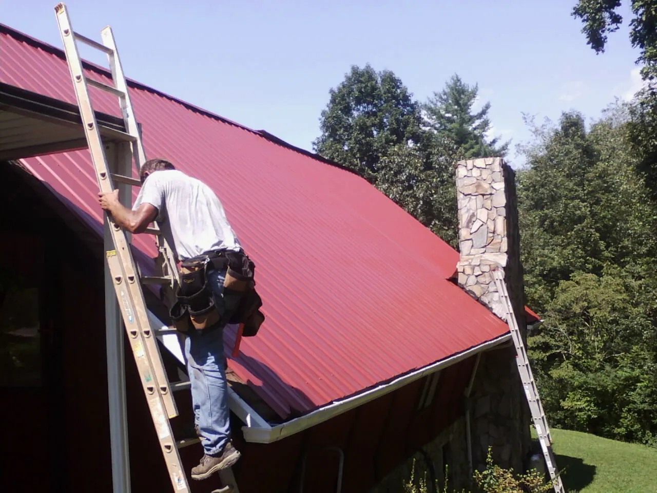 Roofer on a ladder beside a red metal roof and stone chimney — Soffit & Fascia Repair by Roofing Brothers in Encampment